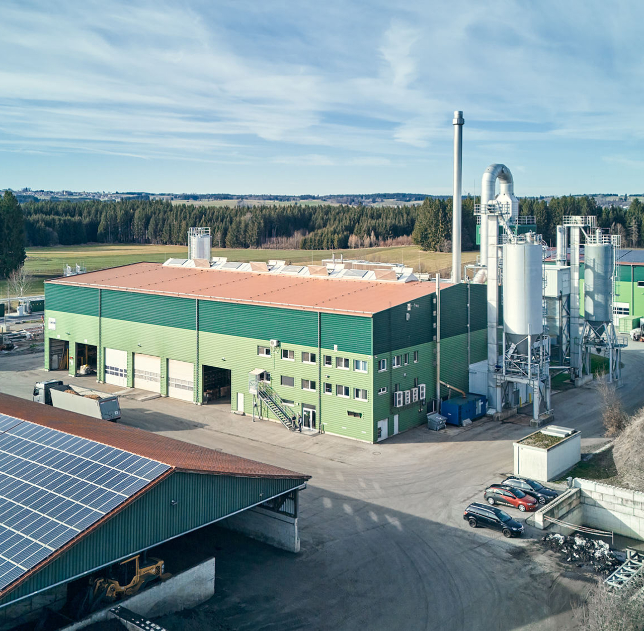 Moderne Produktionsanlage mit Solarpanelen und silbernen Silos, umgeben von Natur und Industrieflächen.