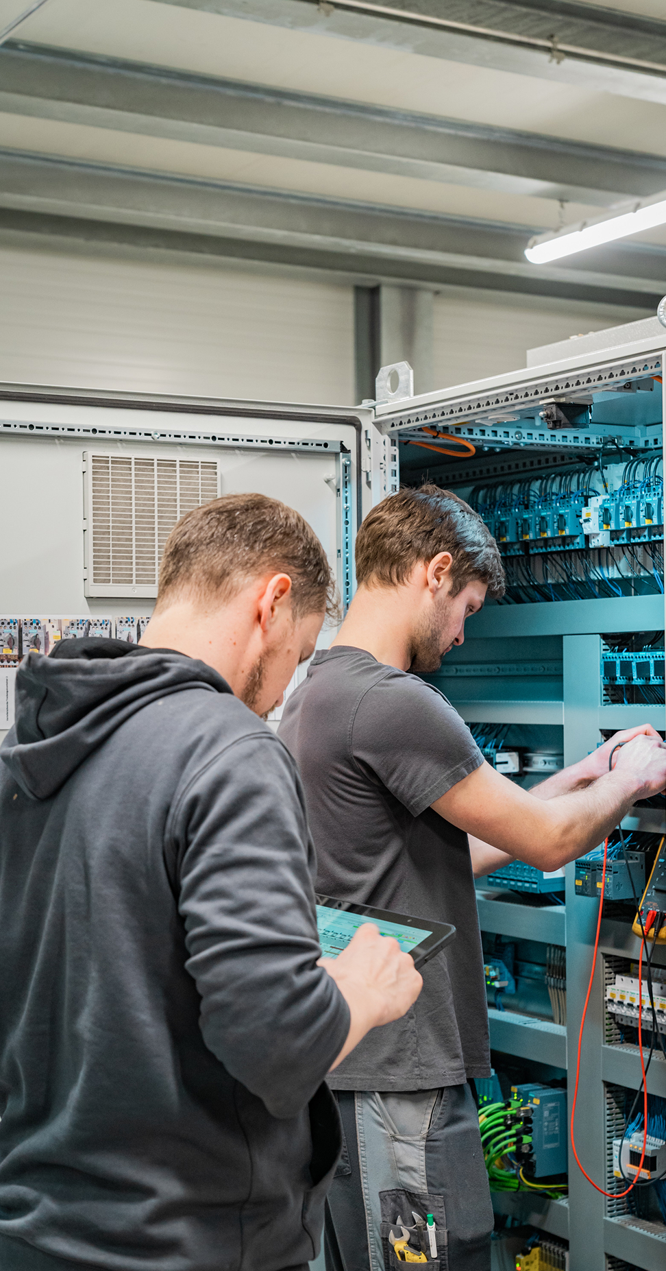 Zwei Techniker arbeiten an einem Schaltschrank und überprüfen elektrische Verbindungen mit einem Tablet in der Hand.