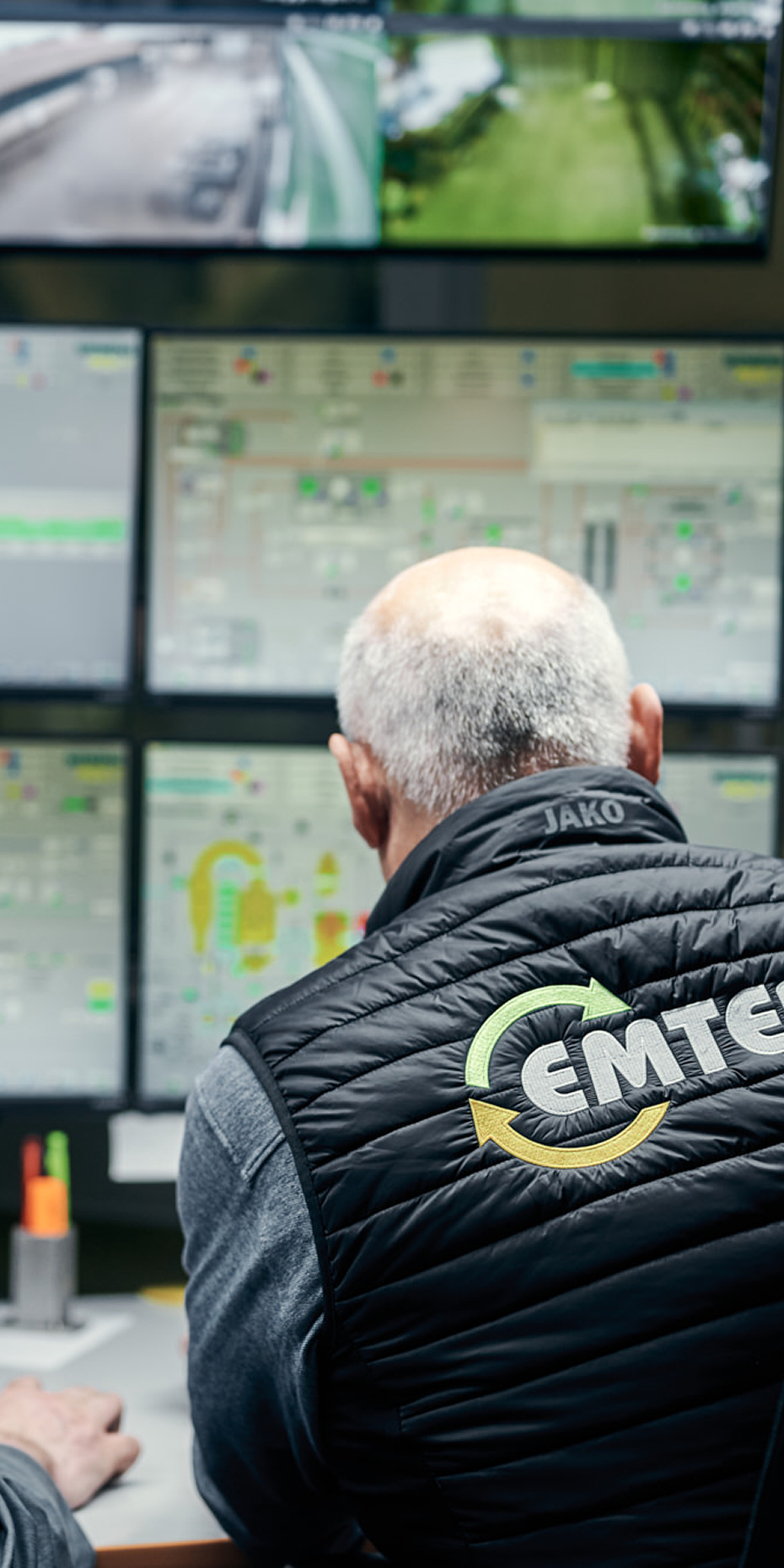 Mitarbeiter, der eine Emter Weste trägt, überwacht mehrere Bildschirme in einem Kontrollraum, um Betriebsabläufe zu steuern und zu optimieren.