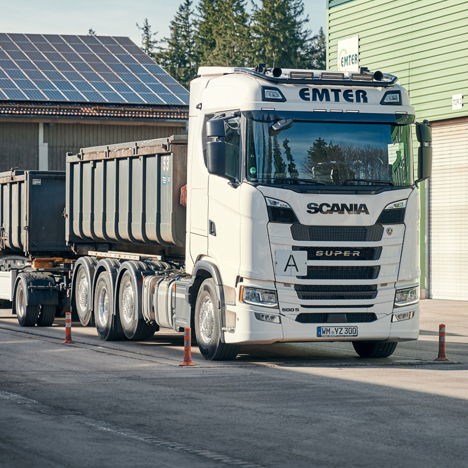Scania Lkw von Emter mit Anhänger vor der Lagerhalle mit Solarzellen auf dem Dach, umgebenden Bäumen im Hintergrund.