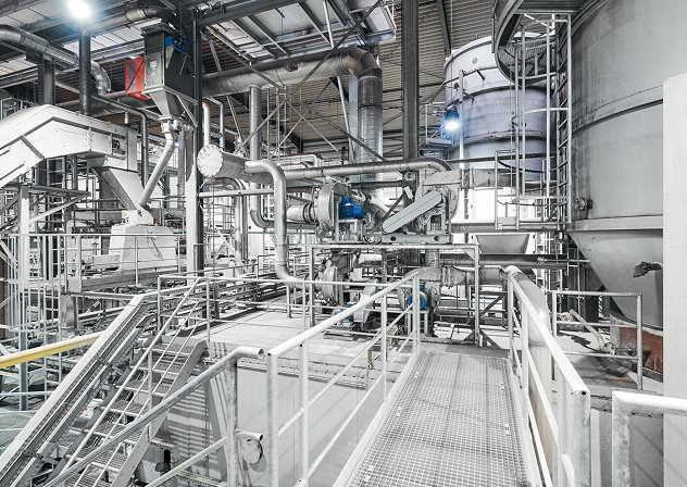 Industrielle Anlage mit Rohren, Maschinen und Treppen, die für die Produktion und Verarbeitung von Materialien genutzt wird.