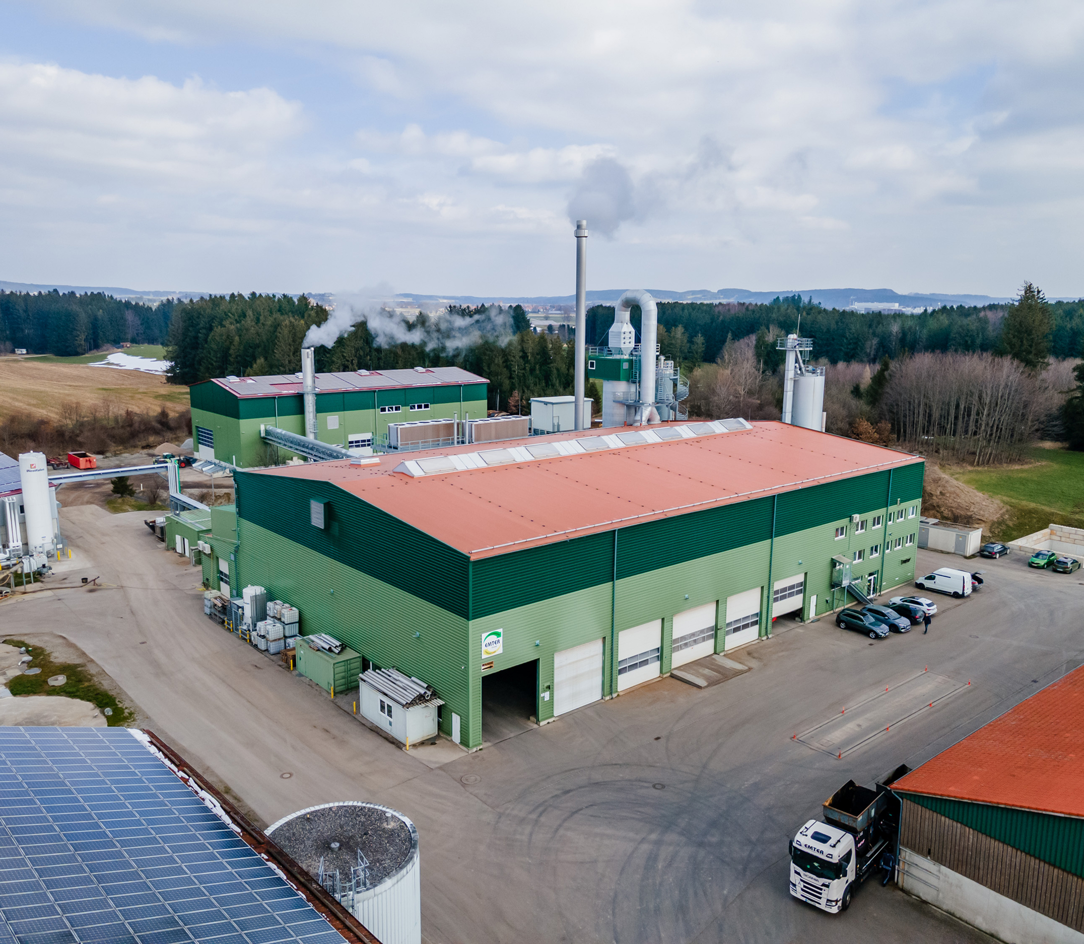 Industriegebäude mit grüner Fassade, Rauchabzügen und Solaranlage, umgeben von Wald und Feldlandschaft.