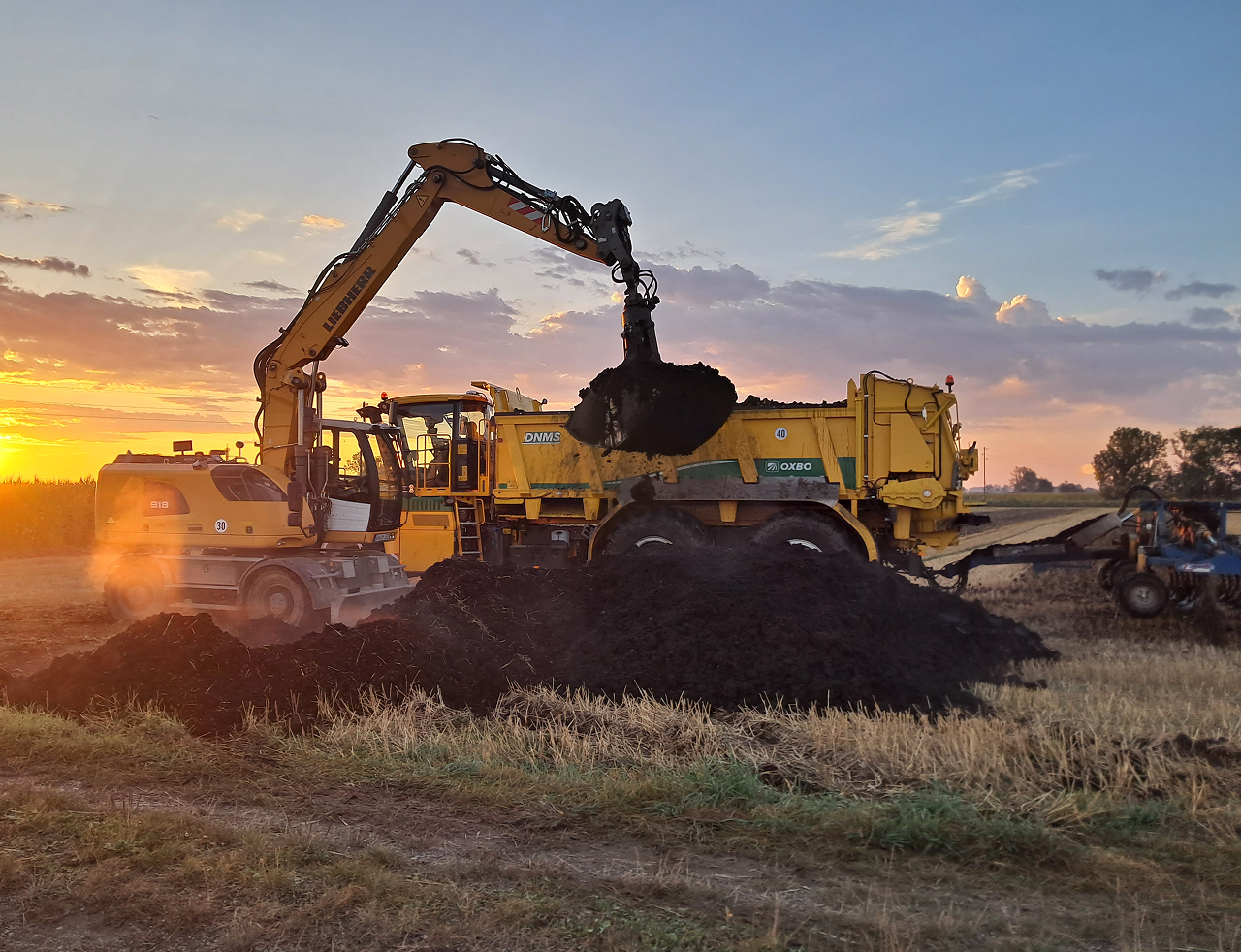 Baugeräte beim Ausbringen von Kompost auf einem Feld bei Sonnenuntergang, umweltfreundliche Landwirtschaftstechnik.